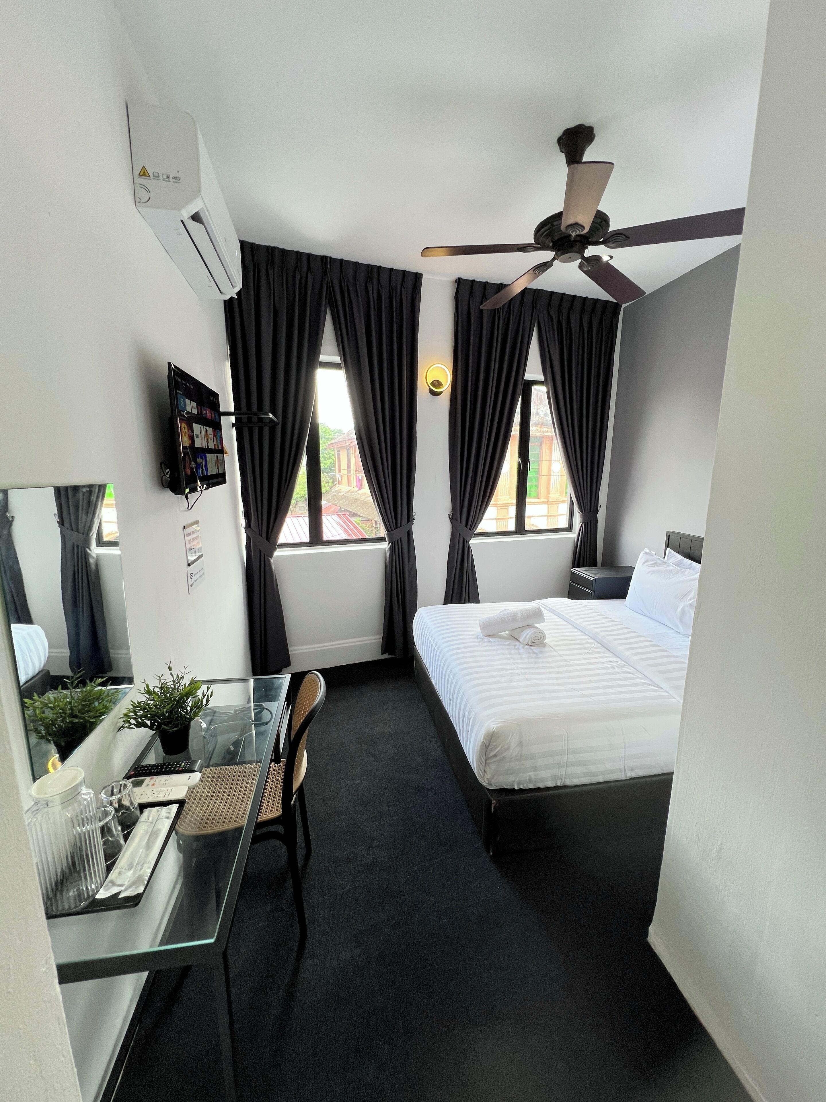 A hotel room with a bed, a desk with a computer, a ceiling fan, and windows with curtains.