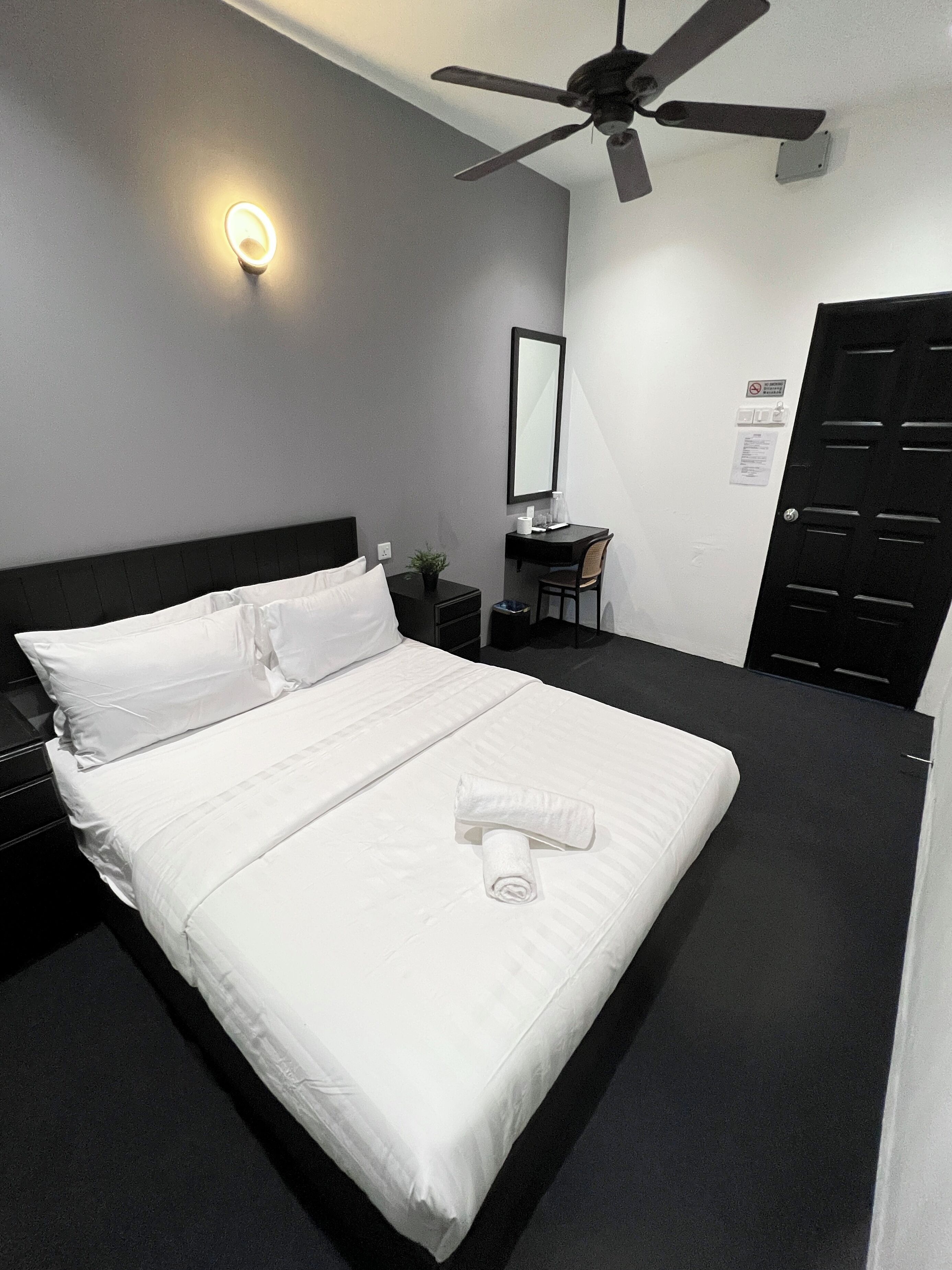 A hotel room with a bed, a desk, a chair, a ceiling fan, and a door.