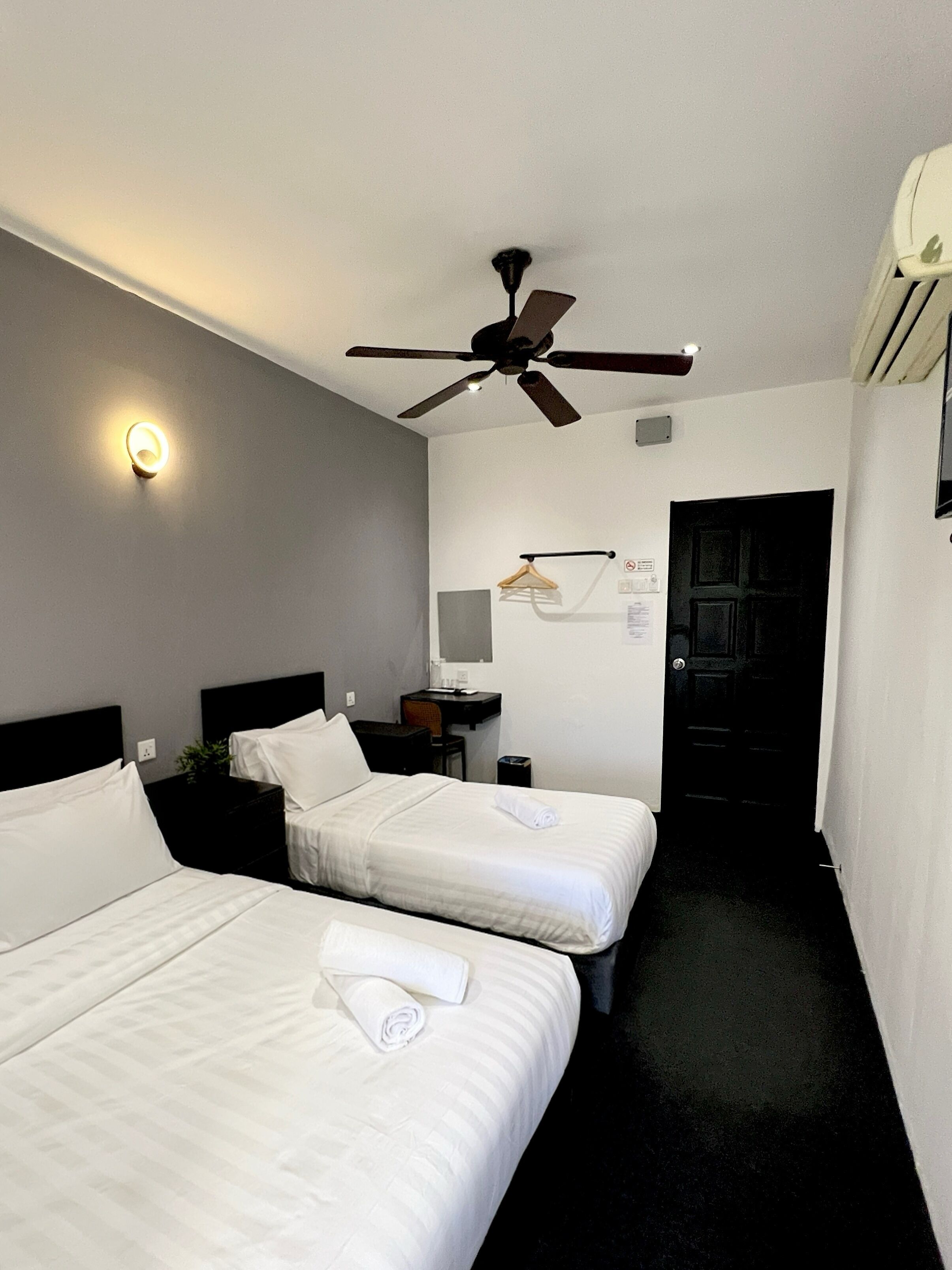 A hotel room with two beds, a ceiling fan, a wall-mounted light, a coat rack, a desk with a chair, and a door.
