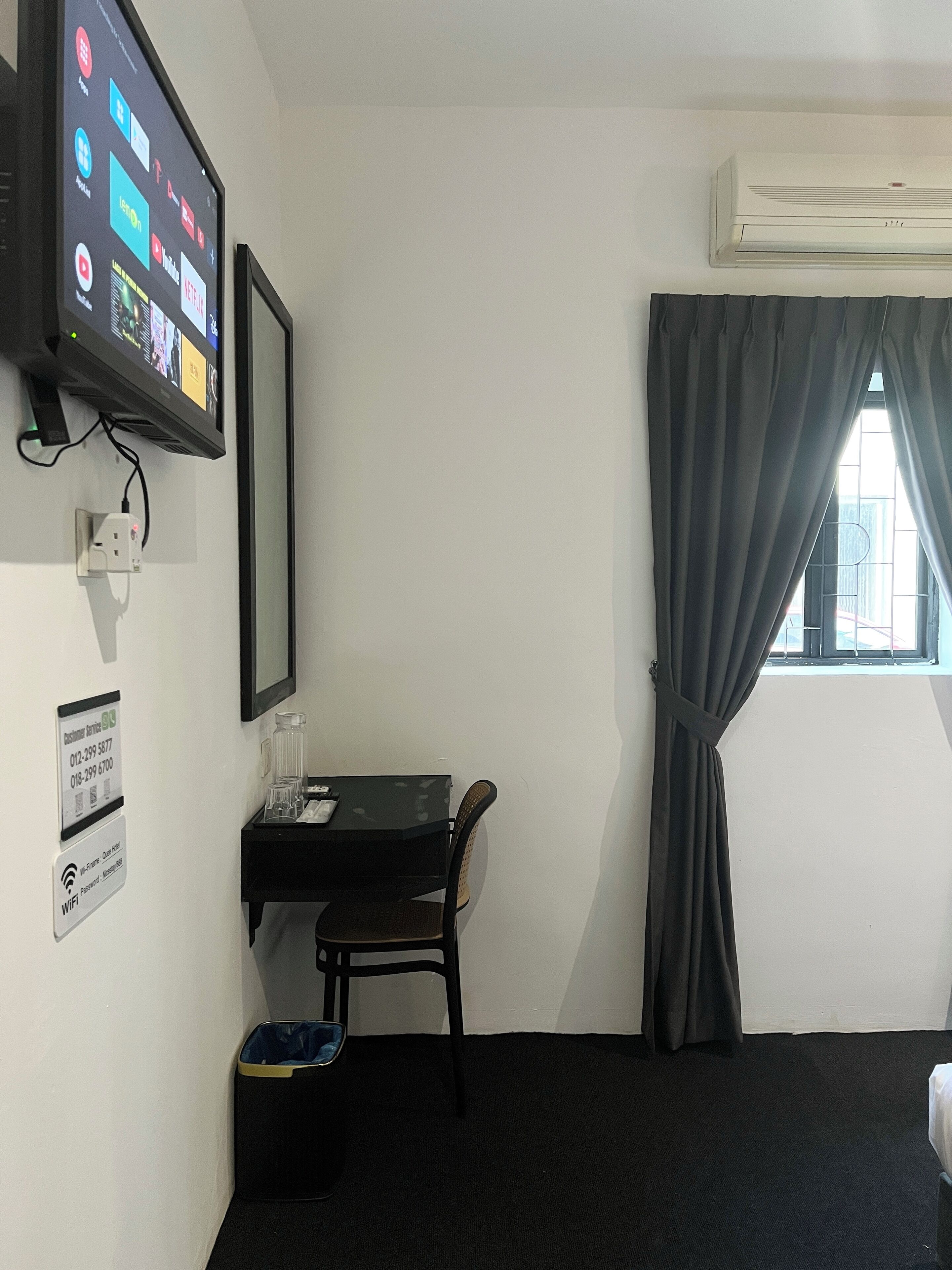 A hotel room with a flat-screen TV mounted on the wall displaying various apps, a desk with a chair, a small trash bin, and a window with curtains.