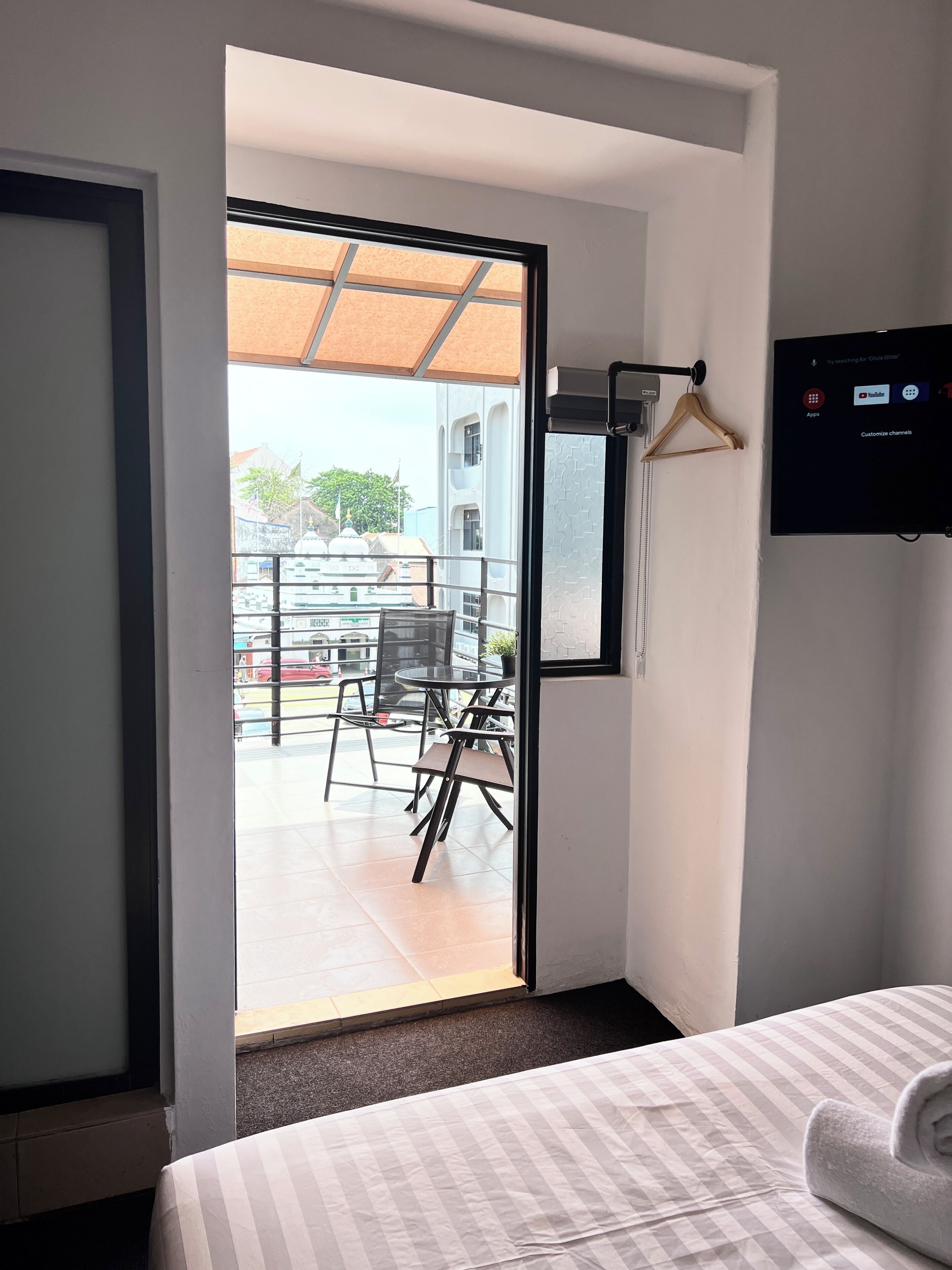 Une chambre d’hôtel comprenant un lit, un téléviseur fixé au mur, une porte-fenêtre coulissante donnant sur un espace extérieur aménagé avec des chaises et une table, ainsi qu’une petite fenêtre.