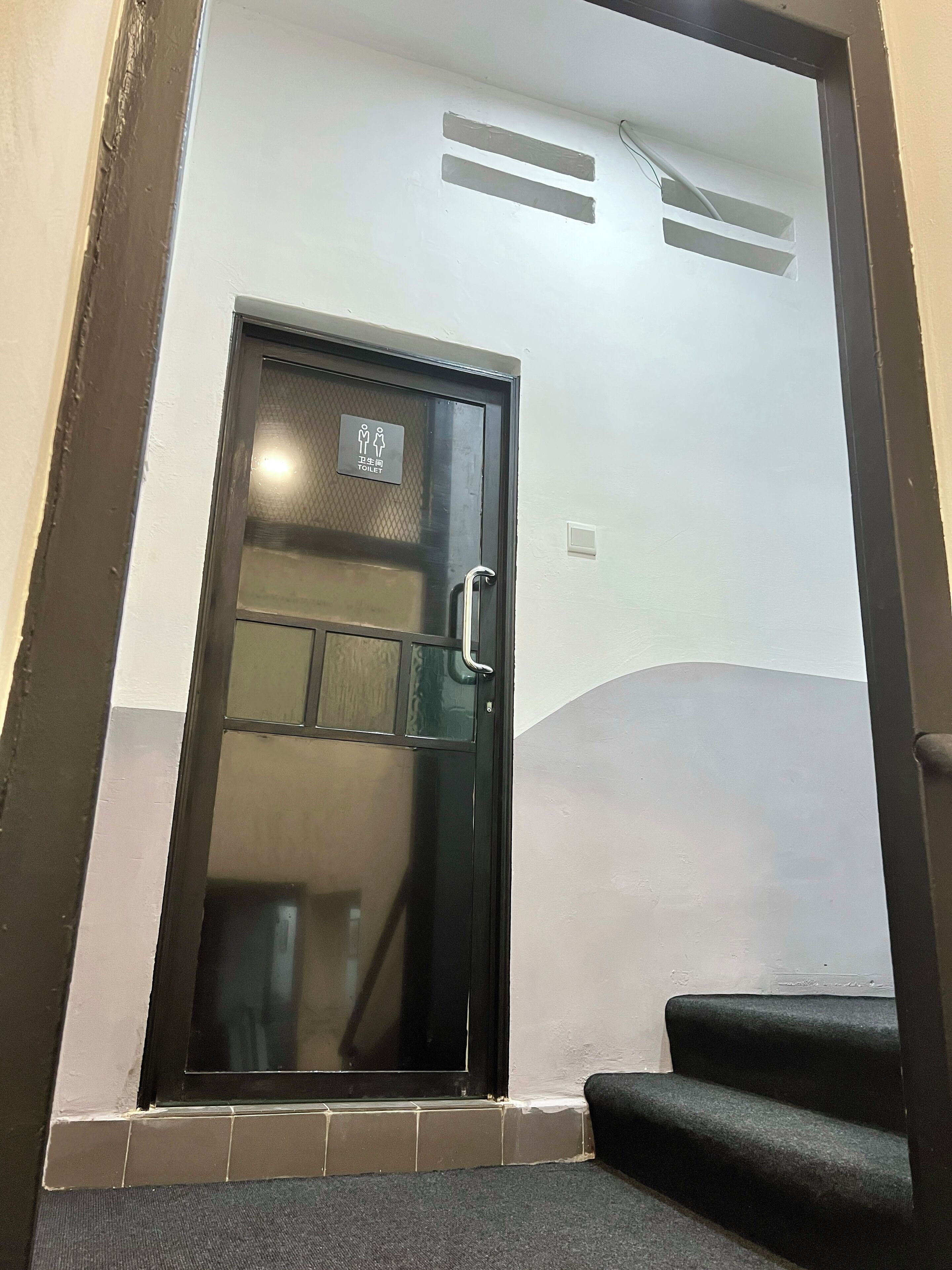 Puerta negra con un panel de vidrio y una barandilla, que da a una zona con baldosas y escalones.