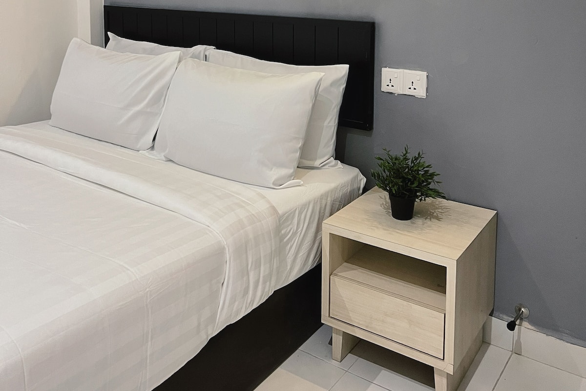 Ein ordentlich bezogenes Bett mit weißen Leinen, ein Nachttisch mit einer Pflanze und eine an der Wand montierte Lampe.
