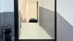A hallway with a black-framed door leading to a room with a white and gray wall, a tiled floor, and a small trash bin to the left. - Quee Hotel (George Town)
