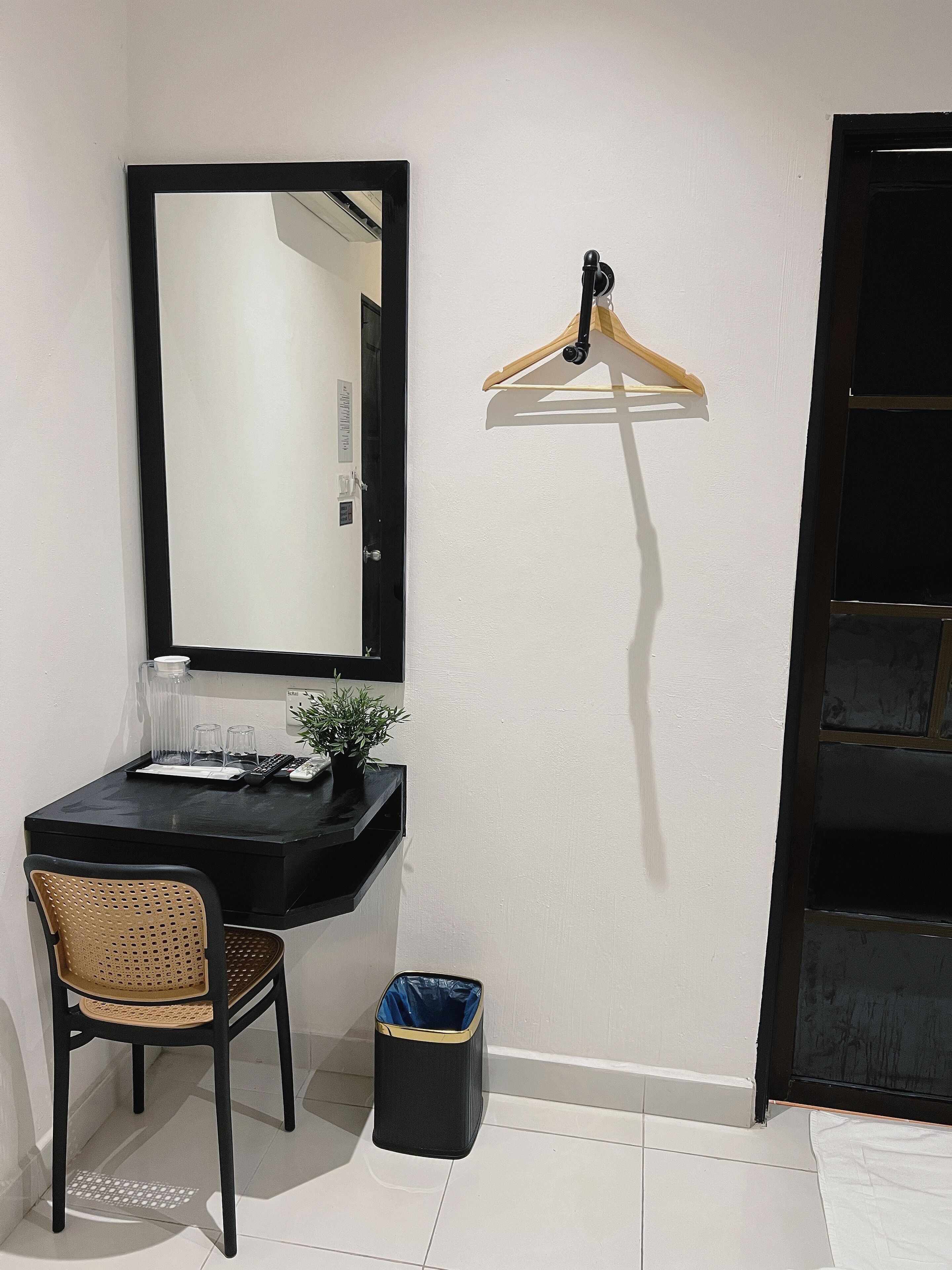 A small room with a black desk, a chair, a trash bin, a hanging clothes rack, and a mirror on the wall.