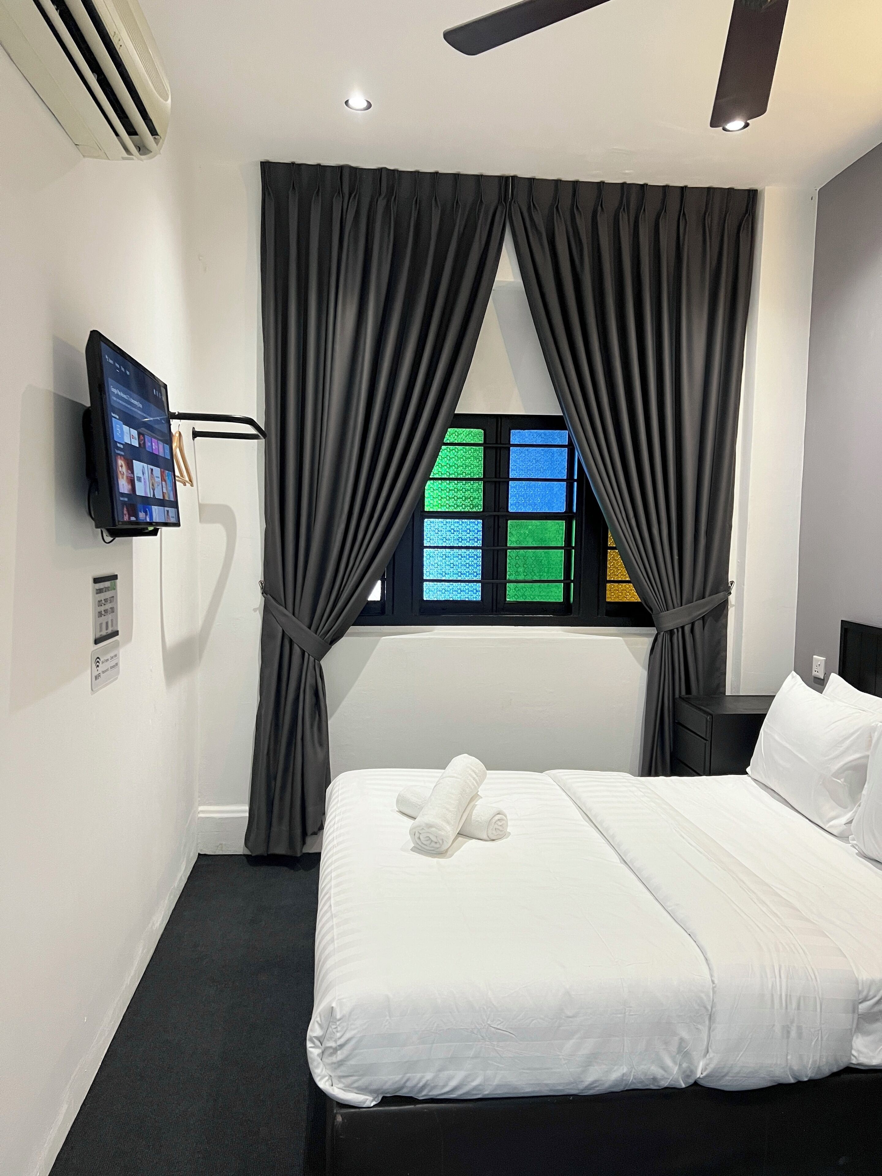 Habitación de hotel con cama, televisor montado en la pared, ventana con cortinas y ventilador de techo.