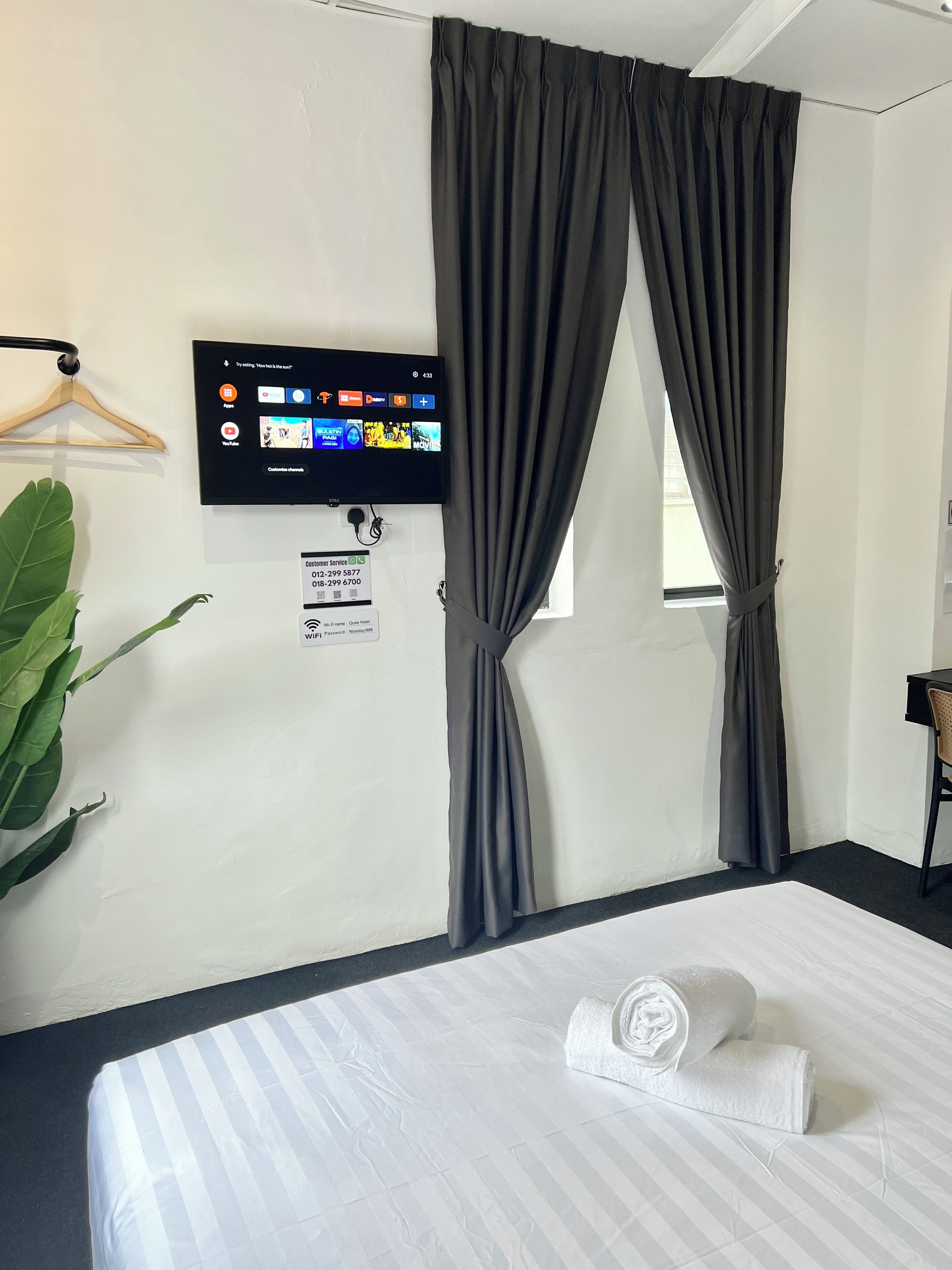 A hotel room with a bed, a TV mounted on the wall, a sign with contact information, and a window with curtains.