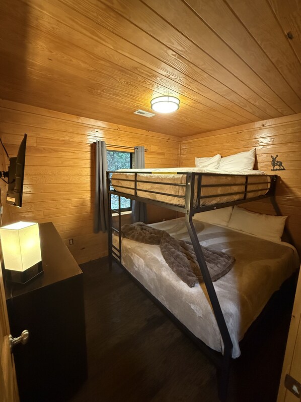 A bunk bed room with wooden walls, a ceiling light, a bedside table with a lamp, and a window with curtains. - Beautiful Cabin w Hot Tub – Sleeps 18 Comfortably (Pigeon Forge)