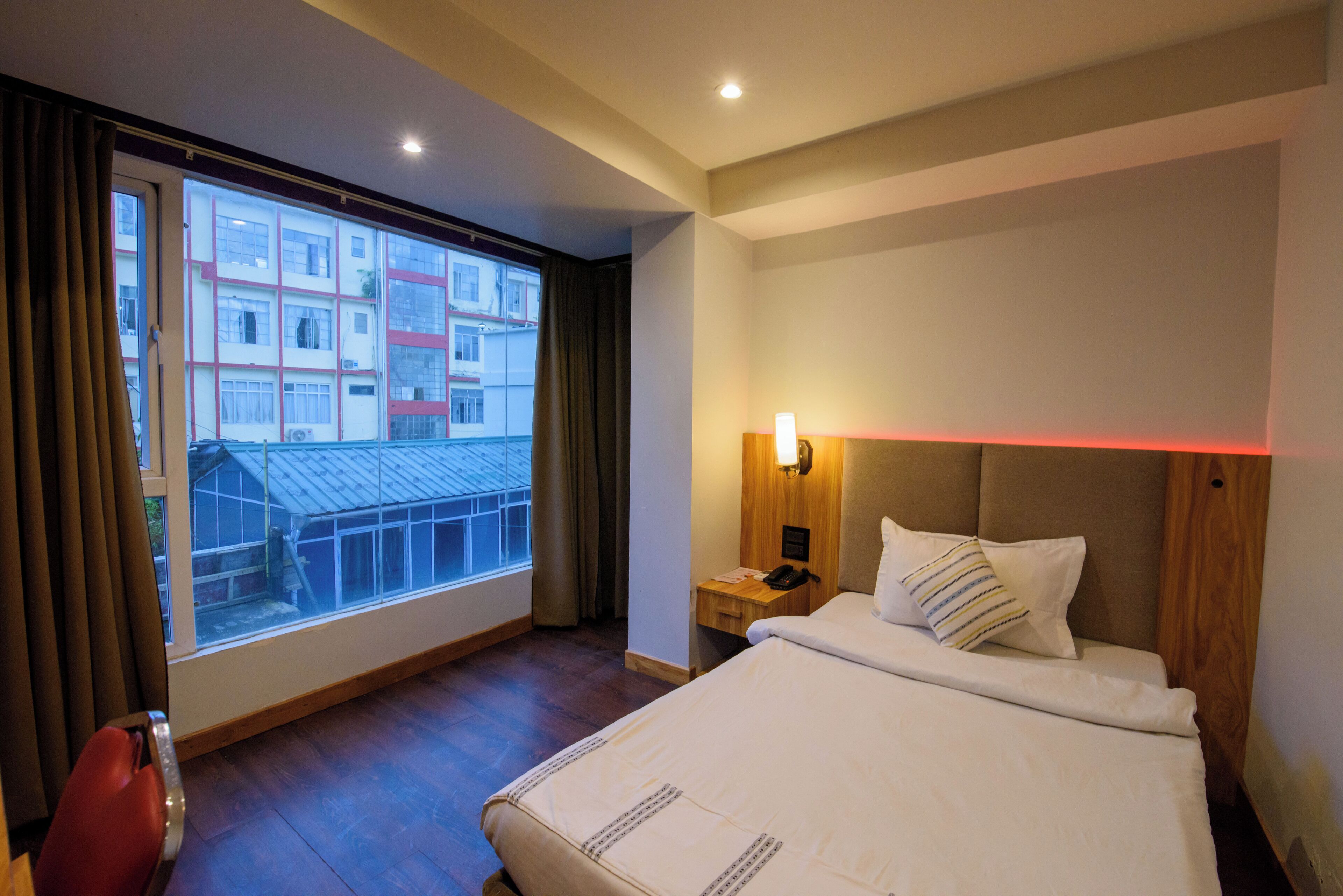 A hotel room with a large window, a bedside table with a lamp, and a bed with pillows.