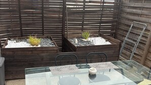 Eine Terrassengaststätte mit einem Tisch aus Glasplatte, schwarzen Stühlen und Holzpflanzgefäßen mit Pflanzen.