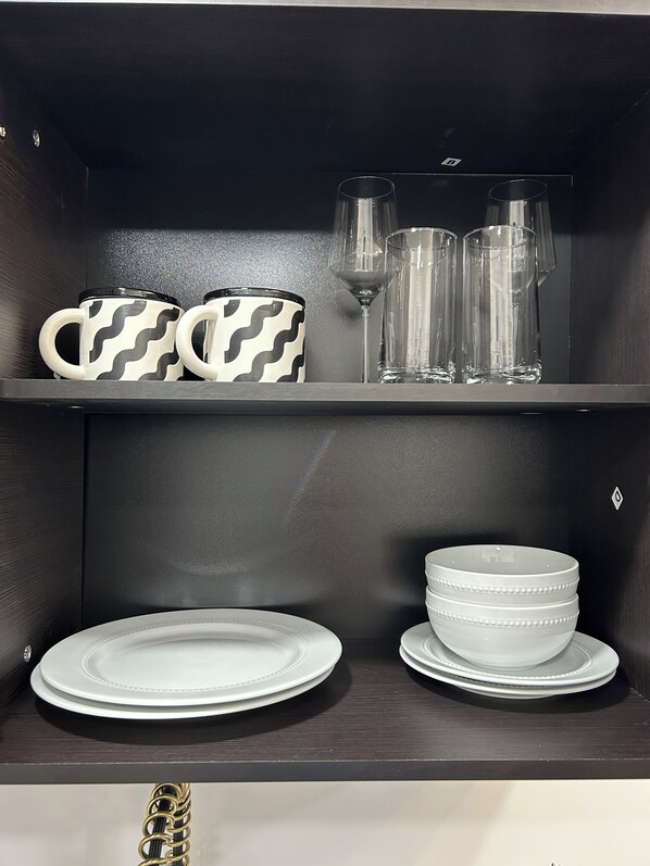 Un estante ordenado con dos tazas estampadas, tres vasos transparentes y dos platos blancos apilados.