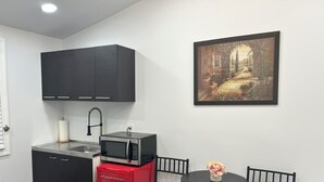Cocina compacta con microondas, un refrigerador pequeño rojo y gabinetes negros. Hay una mesa de comedor con sillas y un solo jarrón con flores. Un cuadro enmarcado cuelga en la pared.
