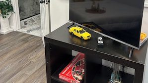 Una habitación con una televisión de pantalla plana grande sobre un soporte negro, un pequeño coche de juguete amarillo, un juego de ajedrez y una maqueta de un paisaje urbano.