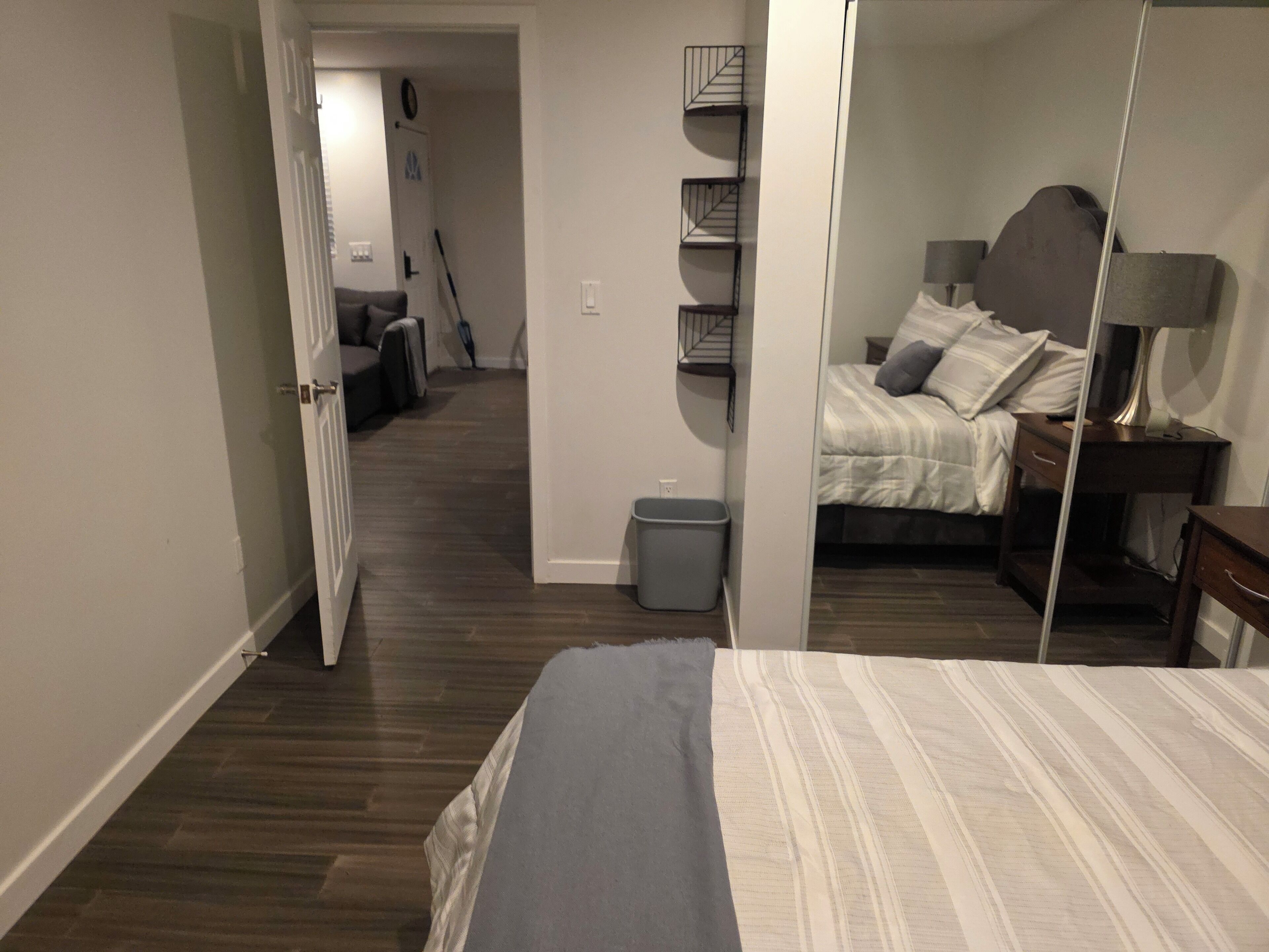 A bedroom with a bed, bedside table, lamp, and a mirror. There is a hallway leading to another room with a couch and a trash bin.