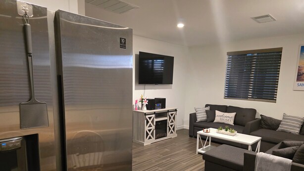 Un salon moderne avec un grand réfrigérateur en acier inoxydable, un téléviseur à écran plat fixé au mur, un canapé gris agrémenté d’un coussin décoratif avec l’inscription « bienvenue à la maison », et une table basse blanche.