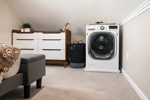 A laundry room with a washing machine, a grey ottoman, a wooden dresser, and a black trash bin. - Cozy Eastwood Guesthouse,private, Convenient, & Comfortable (Syracuse)