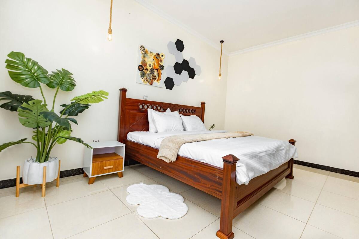 A neatly arranged bedroom with a wooden bed, a nightstand, a large potted plant, and a wall art piece.