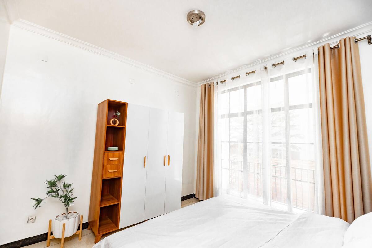 A hotel room with a large bed, a wardrobe, a small potted plant, and a window with curtains.