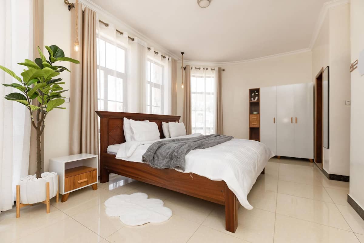 A bedroom with a wooden bed, white curtains, a large potted plant, a bedside table, and a wardrobe.