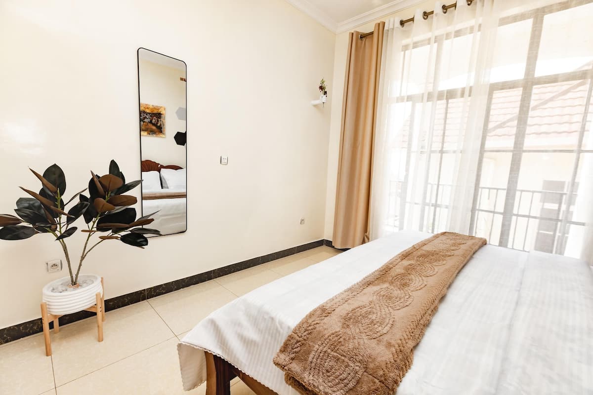 A hotel room with a bed, a mirror, a potted plant, and a window with curtains.