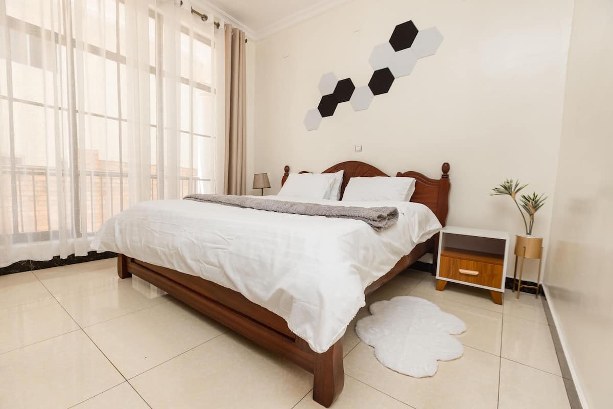 A neatly made bed with a wooden frame and white bedding, a bedside table with a lamp, and a wall-mounted hexagonal decoration.