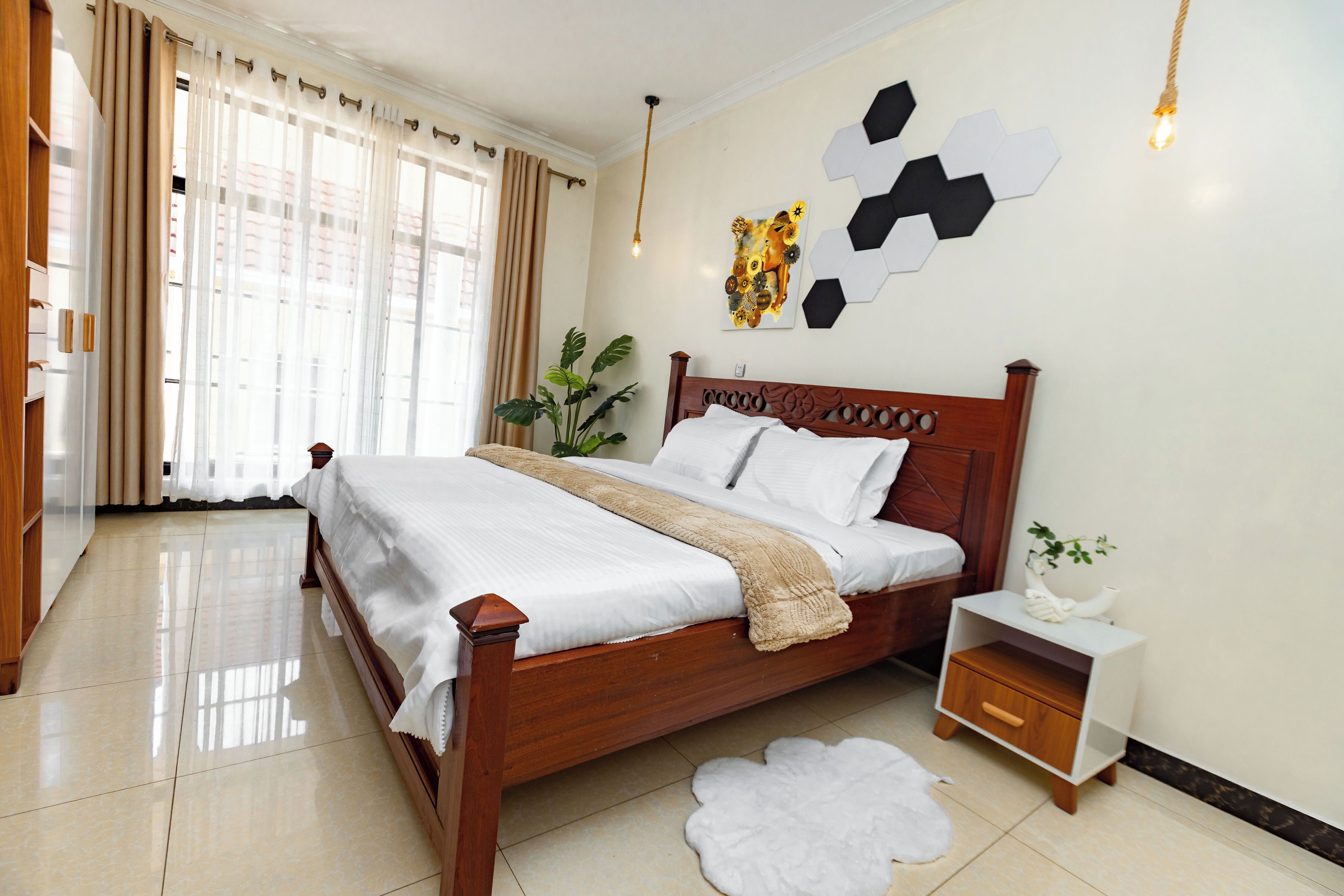 A neatly made bed with a wooden headboard, a bedside table with a drawer, a potted plant, and a wall art piece.