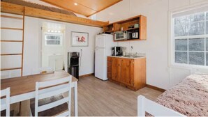 A compact kitchen with wooden cabinets, a microwave, and a refrigerator. There is a dining table with chairs and a window providing a view of the outdoors. - NEW, Promo Pricing - Luxury Woodland Loft Retreat @ Red River Gorge (Stanton)