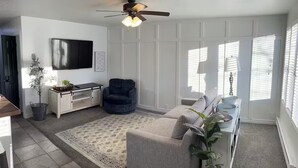 A living room with a sofa, a dark armchair, a TV, a ceiling fan, and a large window with blinds.