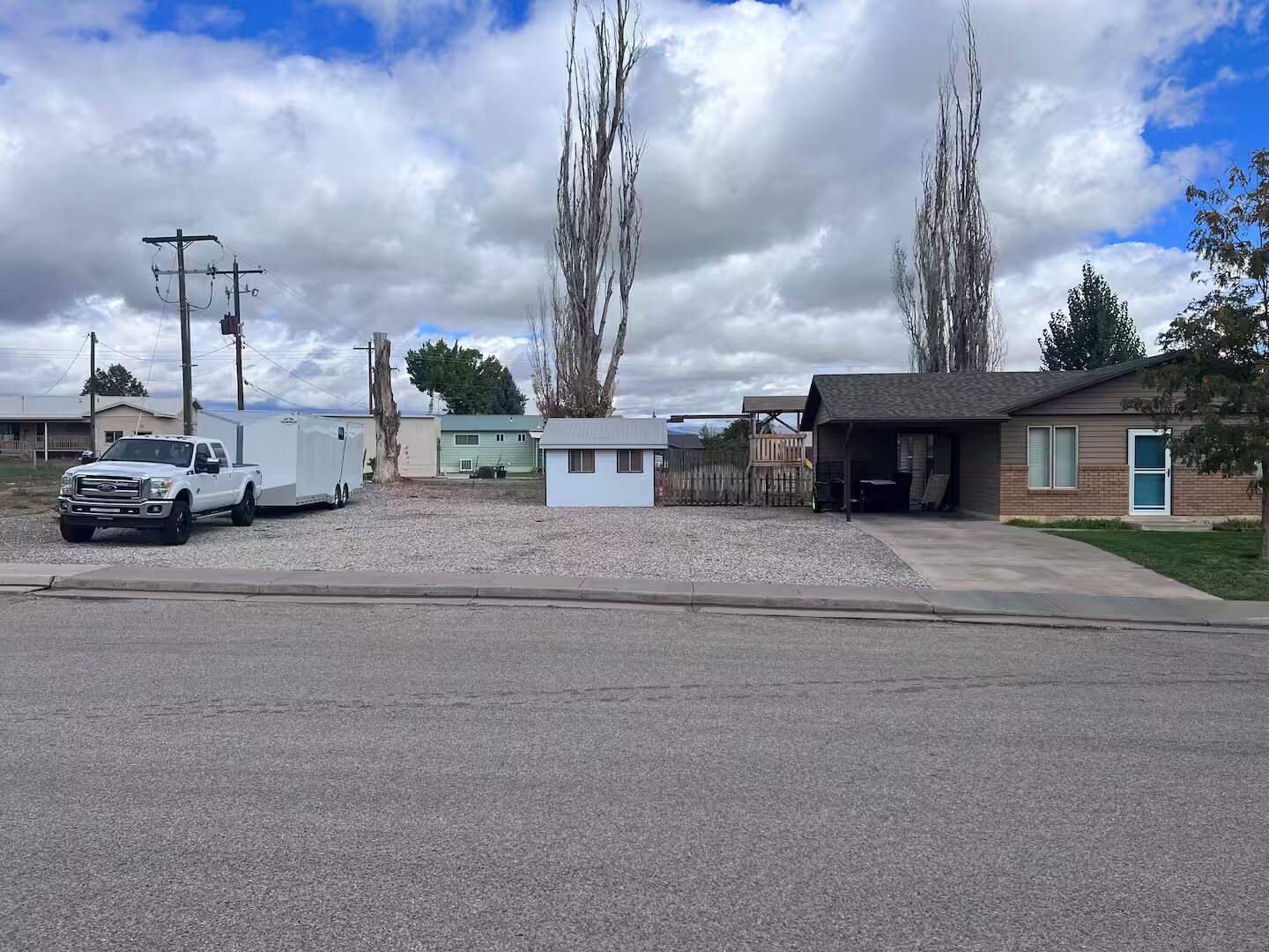 A residential area with a white pickup truck parked on a gravel driveway, a small house with a garage, and a fenced yard.