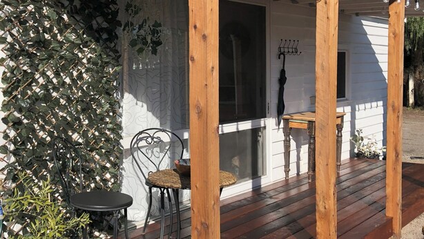 Un porche de madera con fachada blanca, una silla de metal negro, una mesita y un paraguas colgante.