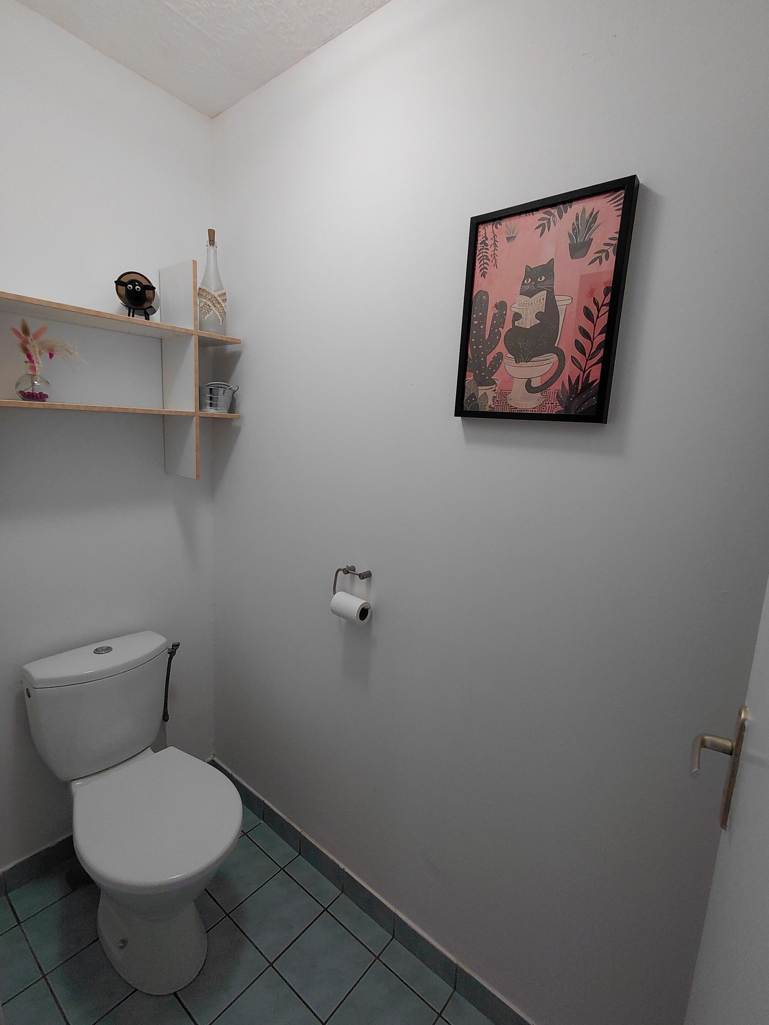 A small bathroom with a toilet, a shelf with decorative items, and a framed picture on the wall.
