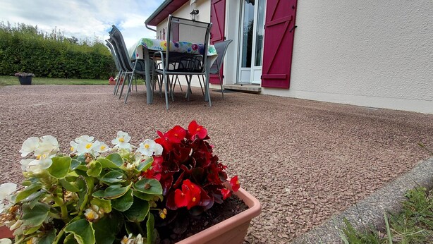 A garden area with a flower bed containing white and red flowers, a patio with a table and chairs, and a building with red shutters and a white door. - House Des 3 filles in cantal 15700 (Ally)