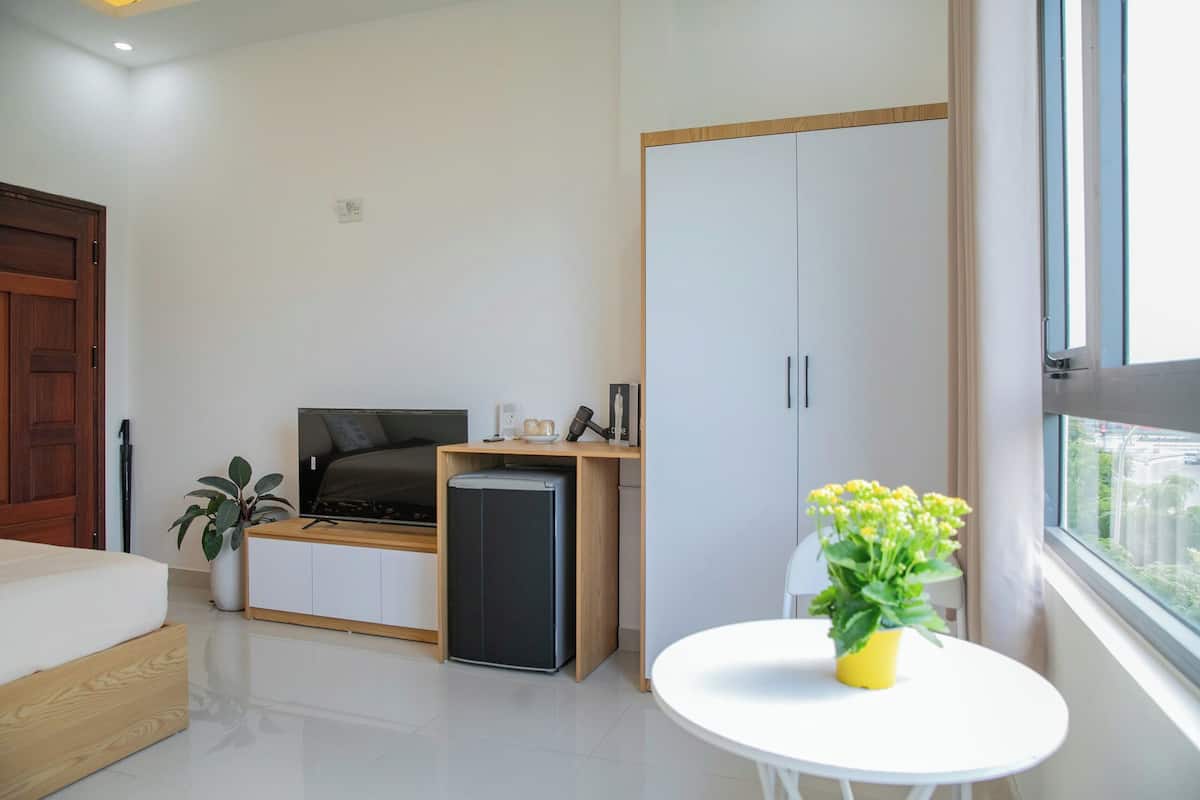 A modern bedroom with a bed, a small desk with a refrigerator, a potted plant, and a window with a view.