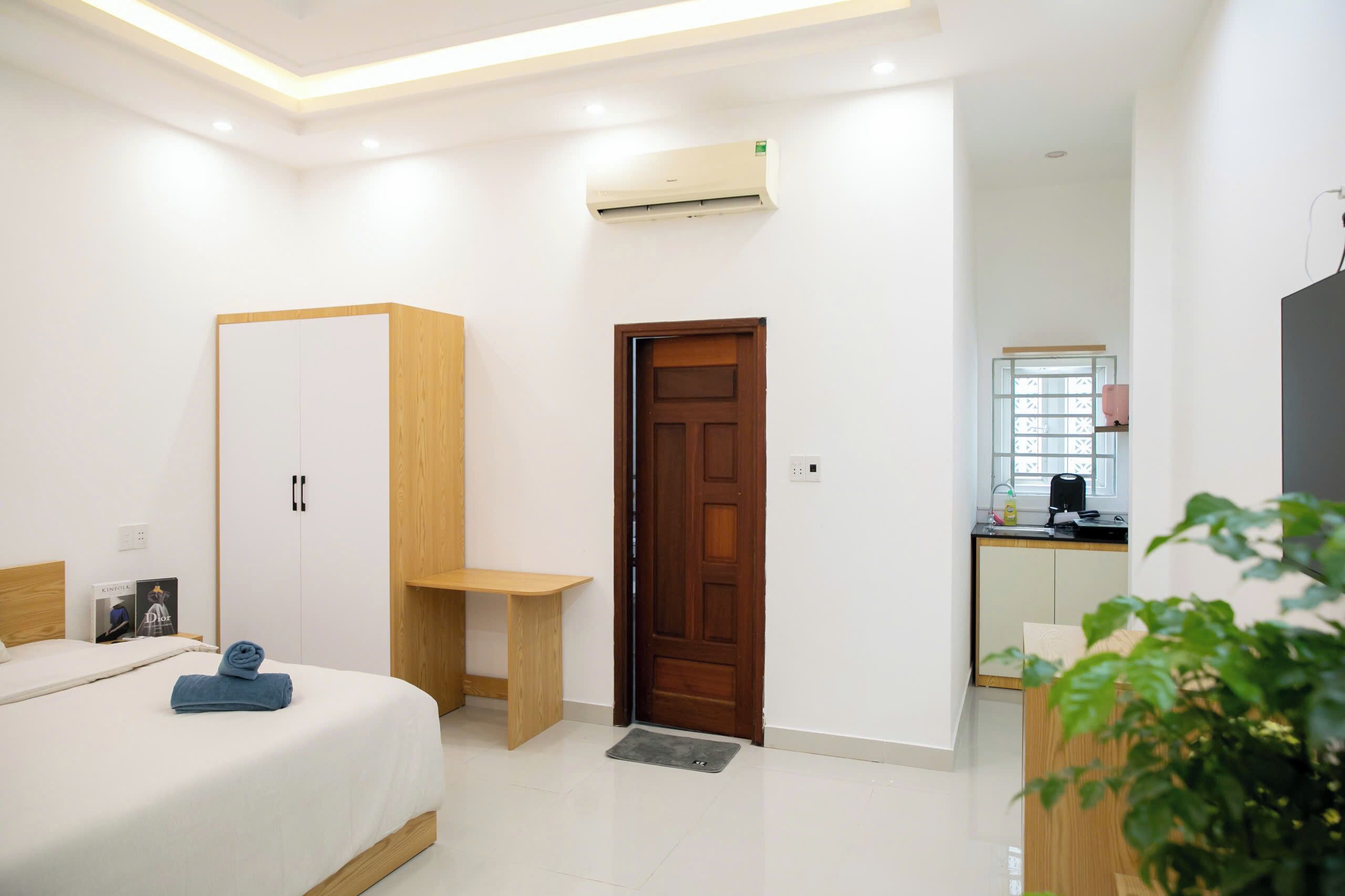 Una habitación de hotel con una cama, un armario de madera, una mesita pequeña, una puerta y una kitchenette al fondo.