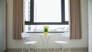 A room with two white chairs and a small round table, a potted plant on the table, and a view of buildings through a large window with beige curtains. - Lavi home (Da Nang)