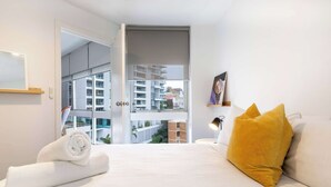 A hotel room with a bed, a yellow pillow, a rolled white towel, a nightstand with a lamp, and a view of buildings through an open door. - Light Filled 1BR  Retreat with Car Park & Pool (Elizabeth Bay)