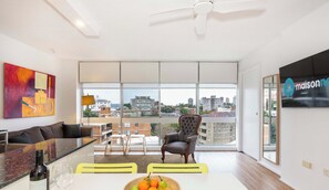 A modern living room with a sofa, a dining table with fruit, a chair, and a city view through large windows.