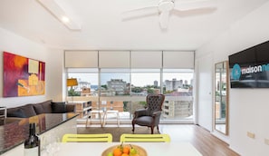 Ein modernes Wohnzimmer mit einer Couch, einem Esstisch mit Obst, einem Sessel und Blick auf die Stadt durch große Fenster.