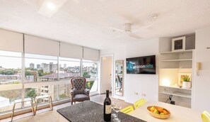 A modern living room with a large window, a flat-screen TV, a dining area with a table and chairs, and a bottle of wine.
