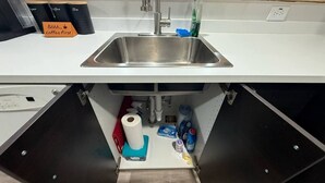 En køkkenvask med en hane i rustfrit stål, et rulle håndklæder og diverse rengøringsartikler under en bordplade.