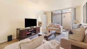 A living room with a flat-screen TV, a wooden cabinet, a sofa, and a coffee table. - Golf View Retreat w/ Sunset Patio, Media Room, Fireplace & Pool/Spa Steps Away (Palm Desert)