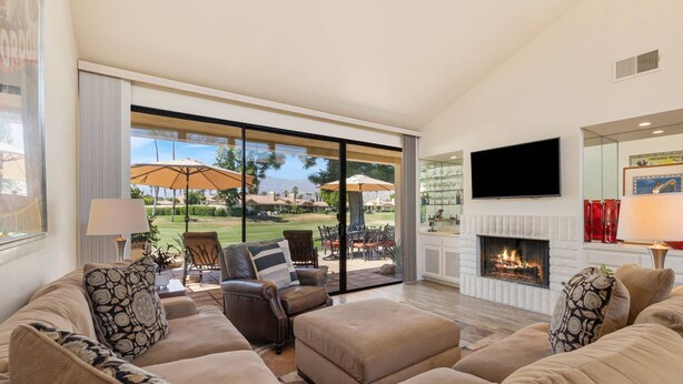 A spacious living room with a large sofa set, a fireplace, and a view of a golf course through a sliding glass door. - Golf View Retreat w/ Sunset Patio, Media Room, Fireplace & Pool/Spa Steps Away (Palm Desert)