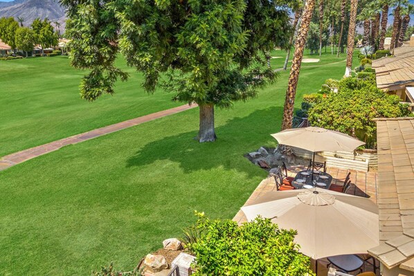 A well-maintained golf course with a clear path, a large tree, and outdoor seating with umbrellas.