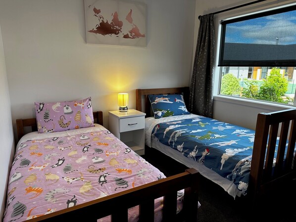 A bedroom with two single beds, a nightstand, a lamp, and a window with a view of greenery.