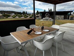 Une terrasse extérieure avec une table en bois et des chaises en osier blanches, une bouteille de champagne et, au fond, une cheminée en pierre.