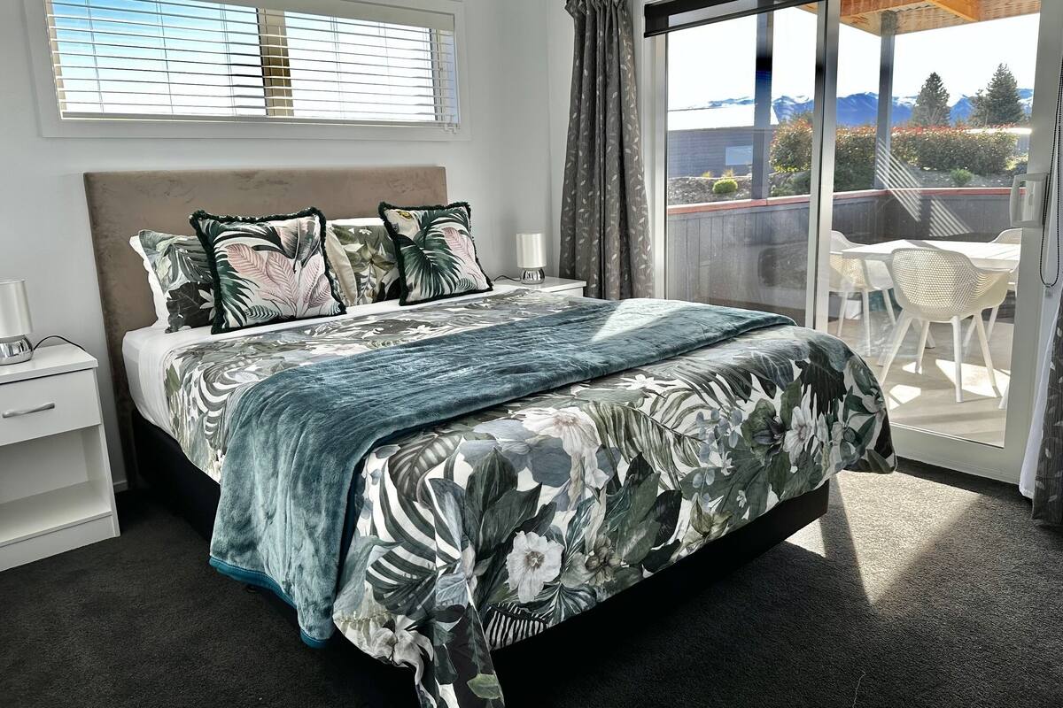 Une chambre avec un grand lit, une table de chevet, une fenêtre avec des stores et une vue sur un balcon aménagé avec du mobilier d’extérieur.