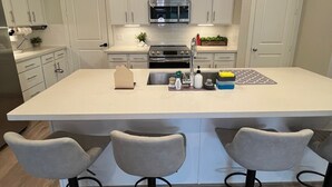 A modern kitchen with white cabinets, a large island with bar stools, stainless steel appliances, and pendant lighting. - Urban Nest HTX (Houston)