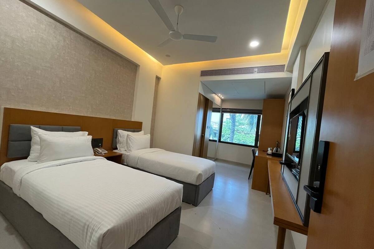 A hotel room with two beds, a ceiling fan, a TV, and a window with a view of greenery.
