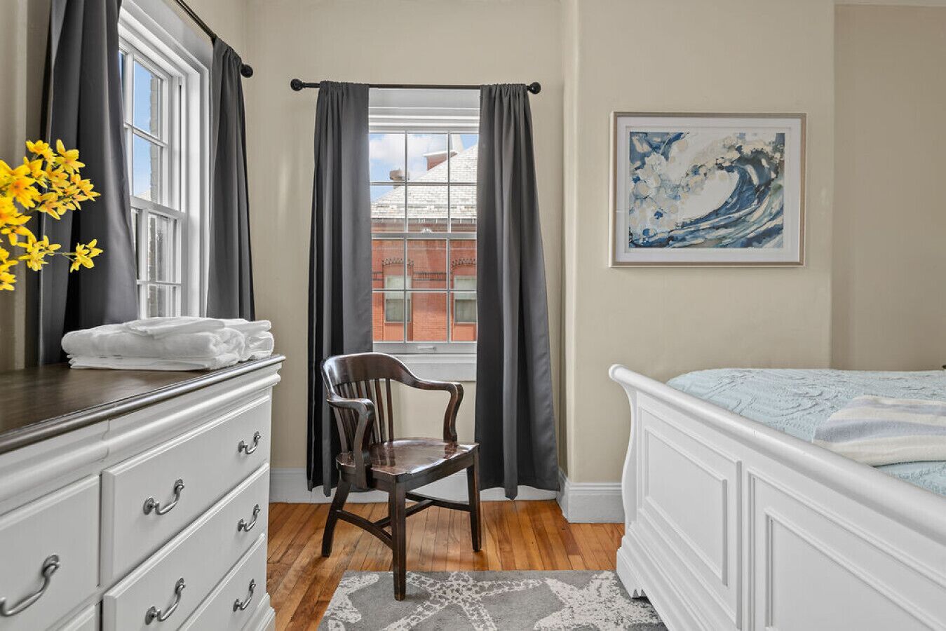 Ein Schlafzimmer mit Bett, Schminktisch, Stuhl, einem Fenster mit Vorhängen und einem Bild an der Wand.