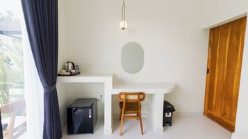 A compact room with a white desk, a wooden chair, a small black refrigerator, a wooden wardrobe, and a hanging light fixture.